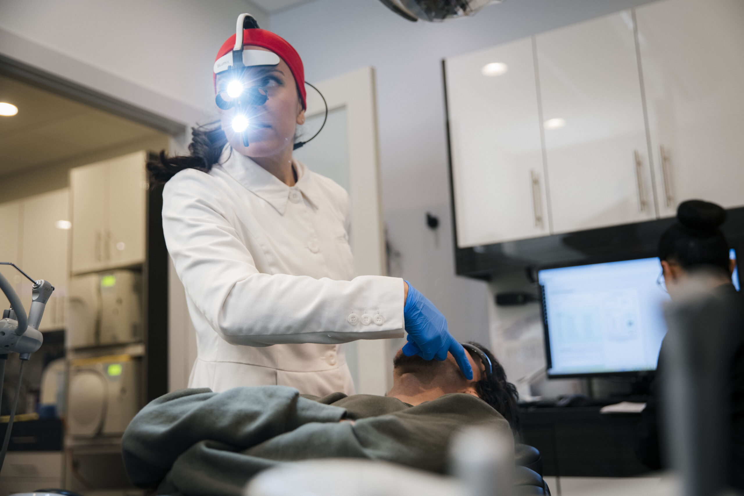 Dentist wearing surgical headlight and red headband performing detailed intraoral procedure on patient.