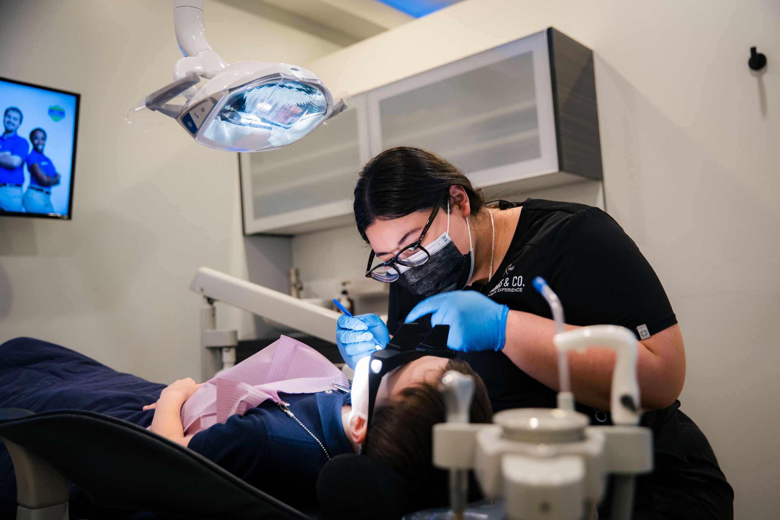 Dentist in black coat and black hair in glasses performing an intraoral procedure on a patient in a modern office
