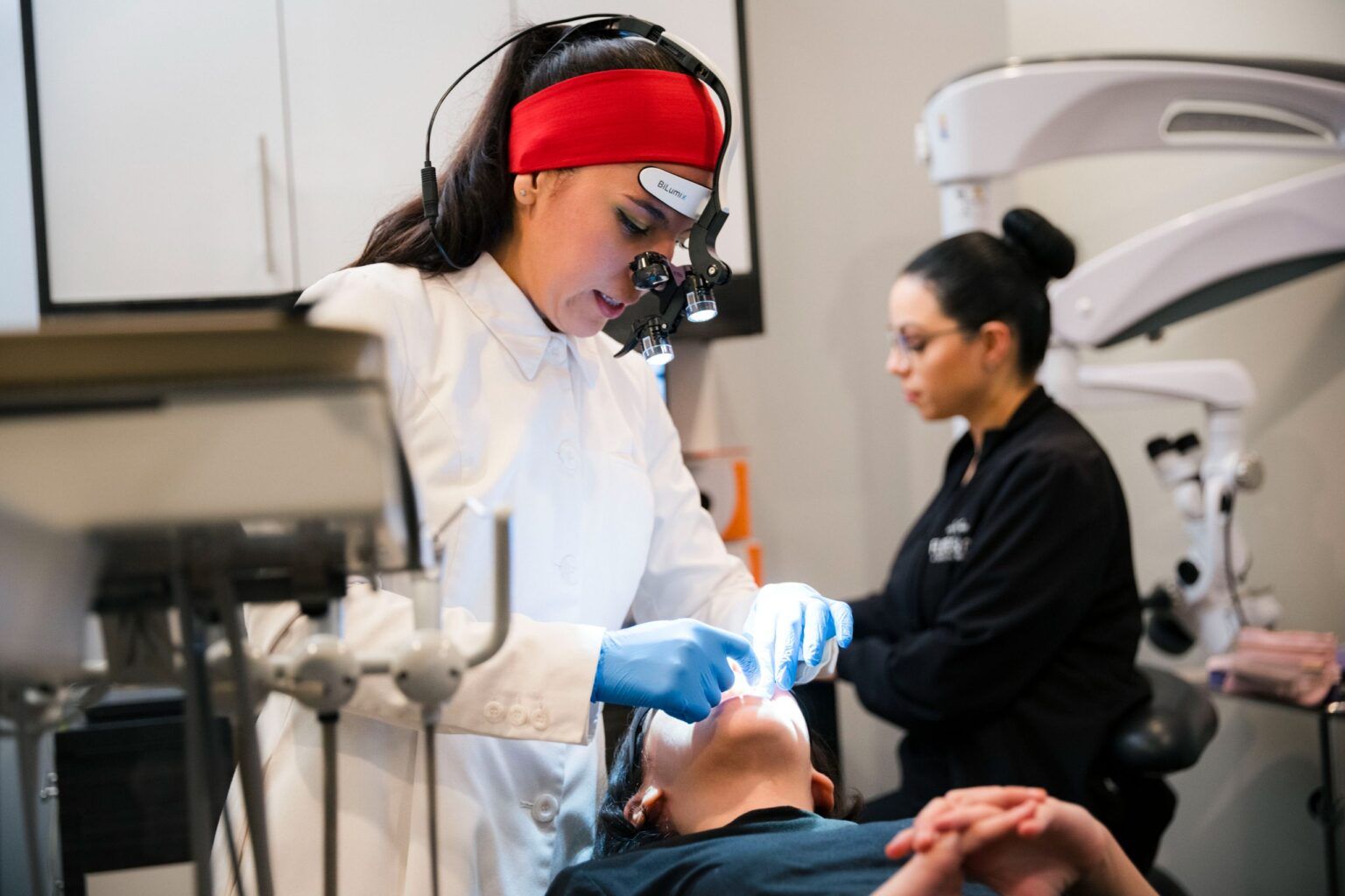 Dentist in white coat and red headband with magnification loupes performing an intraoral procedure on a patient in a modern office