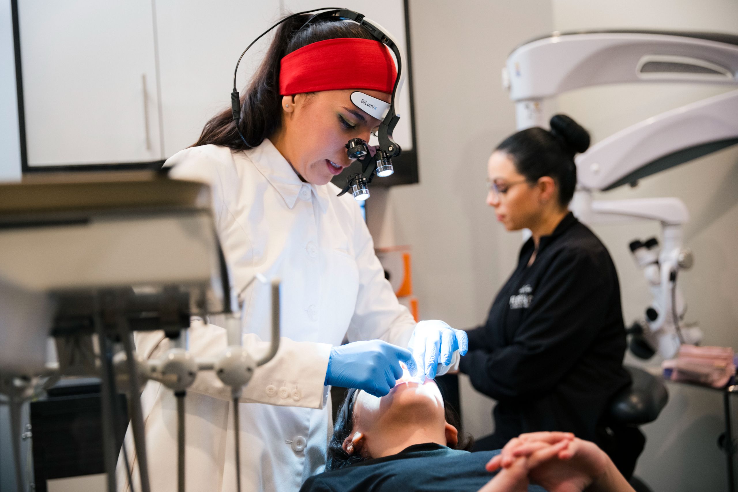 Dentist in white coat and red headband with magnification loupes performing an intraoral procedure on a patient in a modern office